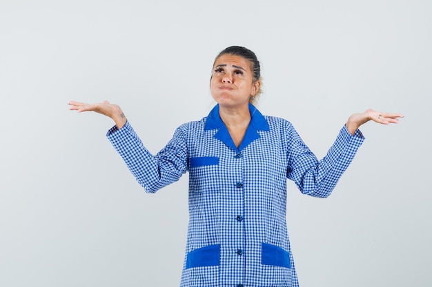 Photo gratuite jeune femme haussant les épaules, les joues gonflées en chemise de pyjama vichy bleu et à la pensif. vue de face.