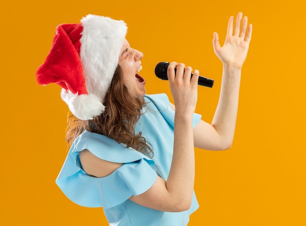 Jeune femme en haut bleu et bonnet de Noel criant au microphone excité fou émotionnel avec bras levé