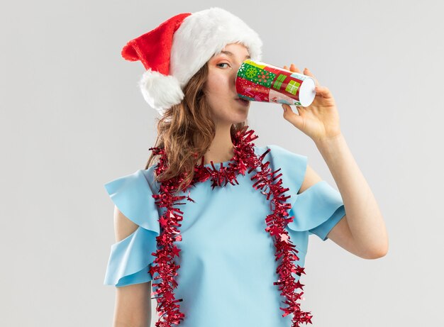 Jeune femme en haut bleu et bonnet de Noel avec des guirlandes autour du cou de boire de la tasse de papier coloré heureux et positif