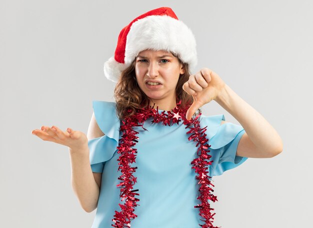 Jeune femme en haut bleu et bonnet de Noel avec des guirlandes autour du cou à être mécontent et confus montrant les pouces vers le bas en levant le bras de mécontentement