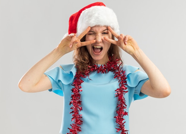 Jeune femme en haut bleu et bonnet de noel avec des guirlandes autour du cou à la folle heureuse et excitée montrant v-sign près des yeux