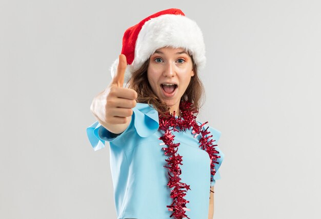 Jeune femme en haut bleu et bonnet de Noel avec des guirlandes autour du cou à heureux et excité montrant les pouces vers le haut