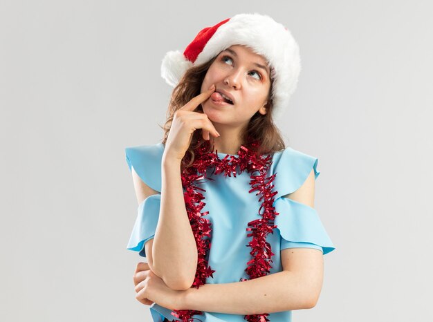 Jeune femme en haut bleu et bonnet de Noel avec guirlandes autour du cou à la perplexité