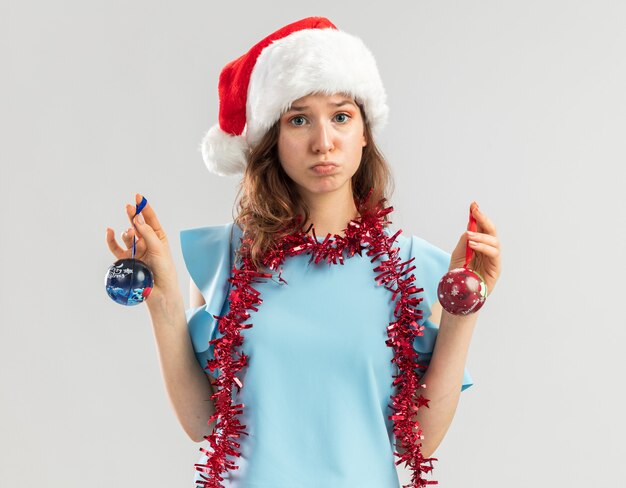 Photo gratuite jeune femme en haut bleu et bonnet de noel avec guirlandes autour du cou tenant des boules de noël à la confusion et mécontent