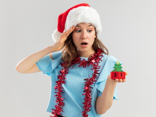 Jeune femme en haut bleu et bonnet de noel avec guirlandes autour de son cou tenant des cubes de jouet avec happy ney year date regardant cubes étonné