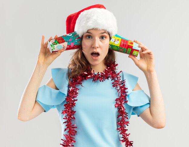 Jeune femme en haut bleu et bonnet de Noel avec guirlandes autour de son cou tenant des gobelets en papier colorés sur ses oreilles à la surprise