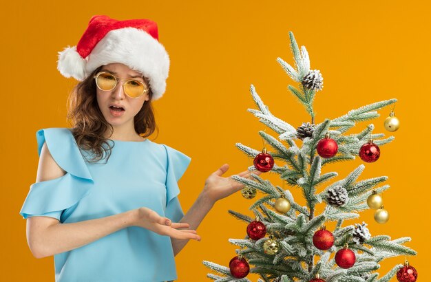 Jeune femme en haut bleu et bonnet de Noel portant des lunettes jaunes debout à côté d'un arbre de Noël présentant les bras des mains l'air confus sur le mur orange