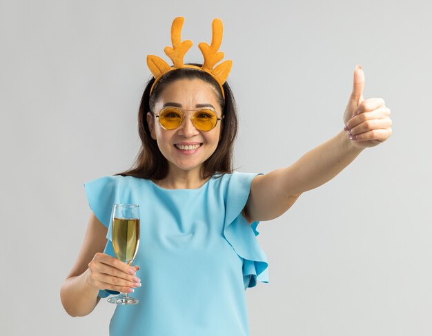 Jeune femme en haut bleu portant une jante drôle avec des cornes de cerf et des verres jaunes tenant un verre de champagne à la recherche de sourire heureux et gai montrant les pouces vers le haut
