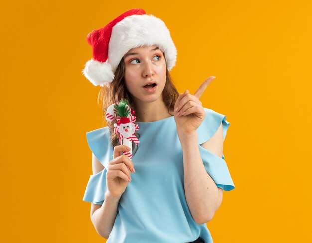 Jeune femme en haut bleu et santa hat holding canne à sucre de Noël à côté surpris en pointant avec l'index à quelque chose