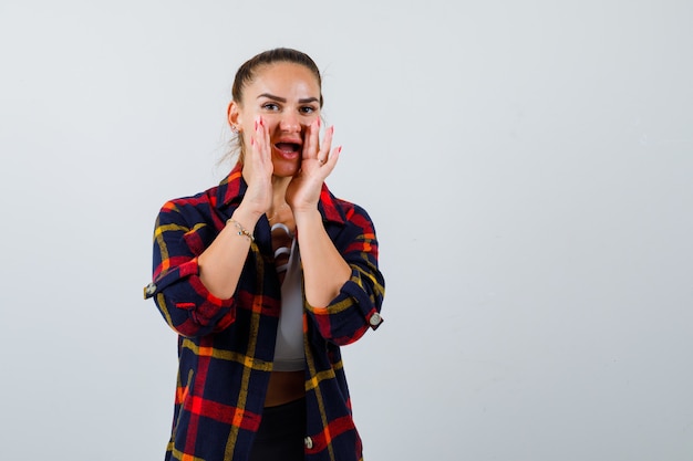 Jeune femme en haut, chemise à carreaux avec les mains près de la bouche pour dire le secret et l'air excité, vue de face.