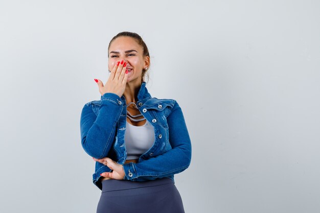 Jeune femme en haut, veste en jean avec la main sur la bouche et l'air heureux, vue de face.