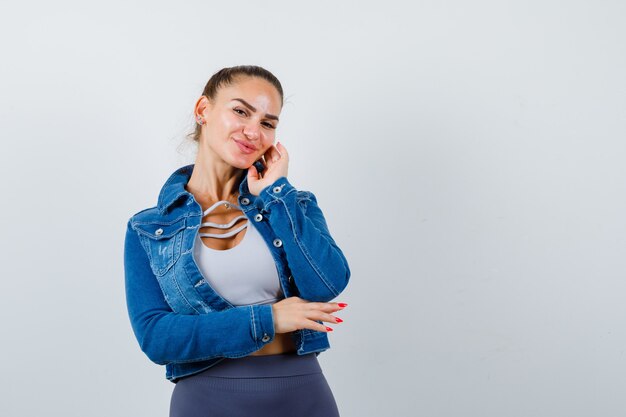 Jeune femme en haut, veste en jean avec la main près de la joue et l'air satisfaite, vue de face.