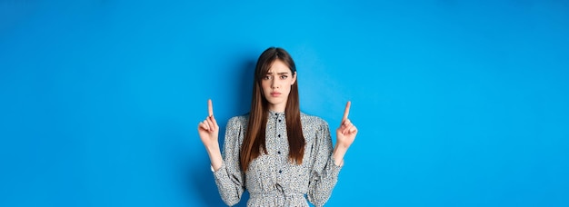 Jeune femme hésitante et inquiète fronçant les doigts pointés vers le haut avec un visage triste debout sur le backgro bleu