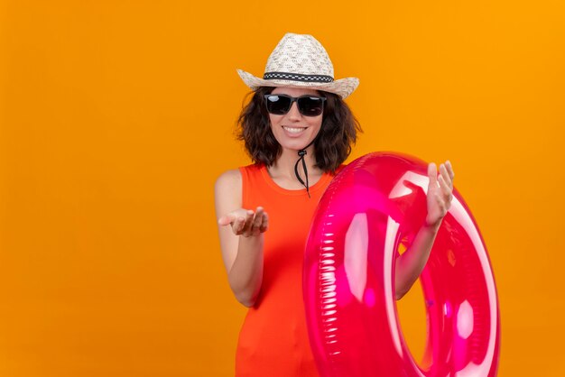 Une jeune femme heureuse aux cheveux courts dans une chemise orange portant un chapeau et des lunettes de soleil tenant un anneau gonflable appelant plus près avec le geste de la main