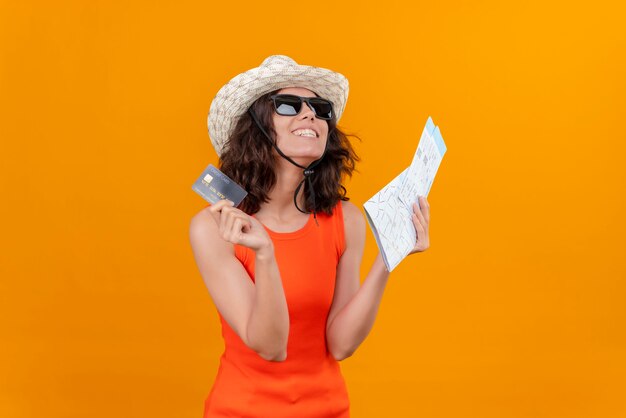 Une jeune femme heureuse aux cheveux courts dans une chemise orange portant un chapeau et des lunettes de soleil tenant une carte et une carte de crédit