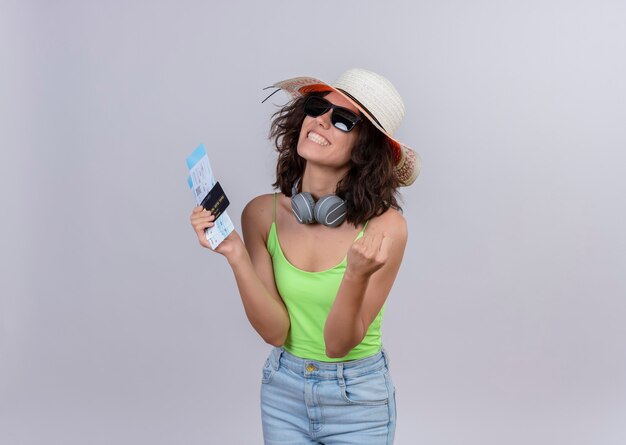 Une jeune femme heureuse aux cheveux courts en vert crop top portant un chapeau de soleil et des lunettes de soleil tenant des billets d'avion et une carte de crédit sur fond blanc