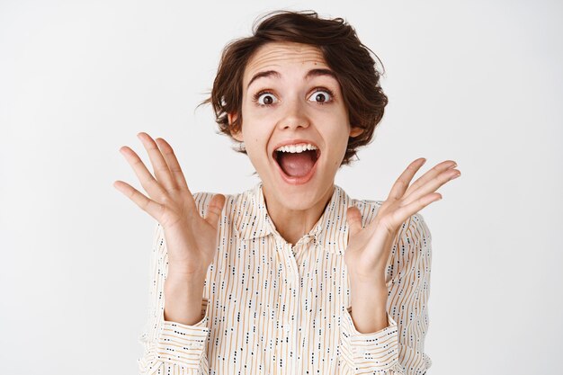 Une jeune femme heureuse crie surprise et émerveillée, semblant étonnée de quelque chose de génial, criant de joie, debout sur un mur blanc