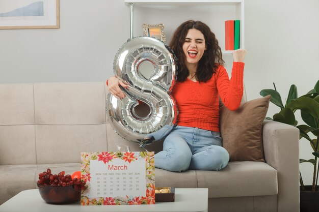 Jeune femme heureuse et excitée en vêtements décontractés souriante joyeusement assise sur un canapé avec un ballon en forme de numéro huit serrant le poing dans un salon lumineux célébrant la journée internationale de la femme le 8 mars