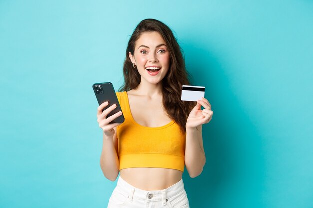 Jeune femme heureuse faisant une commande en ligne, faisant des achats avec une carte de crédit et un smartphone, regarde émerveillée par la caméra, debout sur fond bleu.