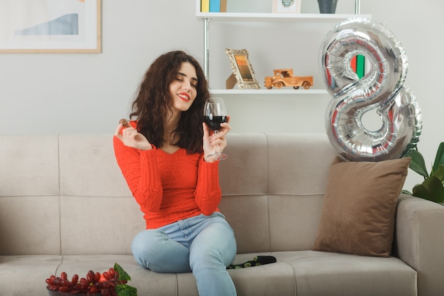Jeune femme heureuse et heureuse en vêtements décontractés souriant joyeusement assise sur un canapé avec un verre de vin et des bonbons au chocolat dans un salon lumineux célébrant la journée internationale de la femme le 8 mars