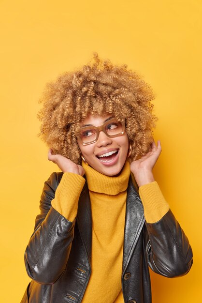 Une jeune femme heureuse montre sa nouvelle coiffure en riant, porte avec joie un pull et une veste en cuir à la mode se sent joyeuse isolée sur fond jaune espace de copie vierge pour votre contenu promotionnel.