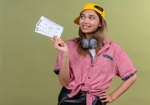 Une jeune femme heureuse portant une chemise rouge et une casquette de baseball jaune avec des écouteurs montrant des billets d'avion tout en regardant côté sur un mur vert