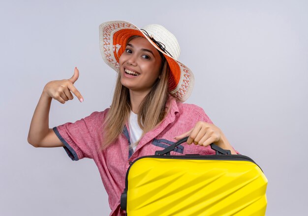 Une jeune femme heureuse portant une chemise rouge pointant sur la valise jaune tout en regardant sur un mur blanc