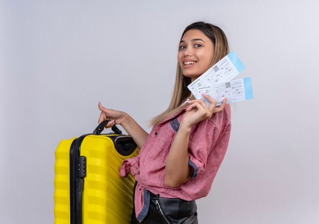 Une jeune femme heureuse portant une chemise rouge tenant une valise jaune avec des billets d'avion tout en regardant sur un mur blanc