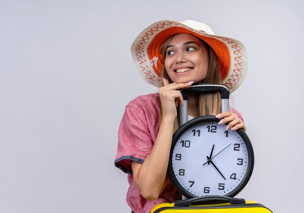 Une jeune femme heureuse portant une chemise rouge tenant une valise jaune avec horloge murale tout en regardant côté sur un mur blanc