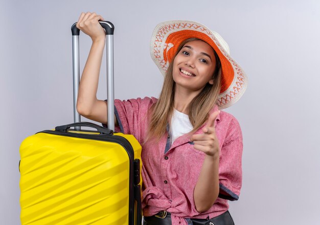 Une jeune femme heureuse portant une chemise rouge tenant une valise jaune tout en pointant sur un mur blanc