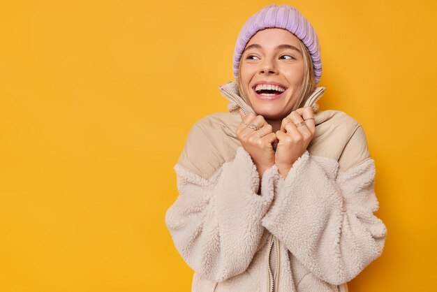 Une jeune femme heureuse porte un bonnet tricoté et une veste chaude détourne le regard souriant concentré loin profite d'une bonne journée d'hiver isolée sur fond jaune avec espace de copie. Fille gaie vêtue de vêtements d'extérieur
