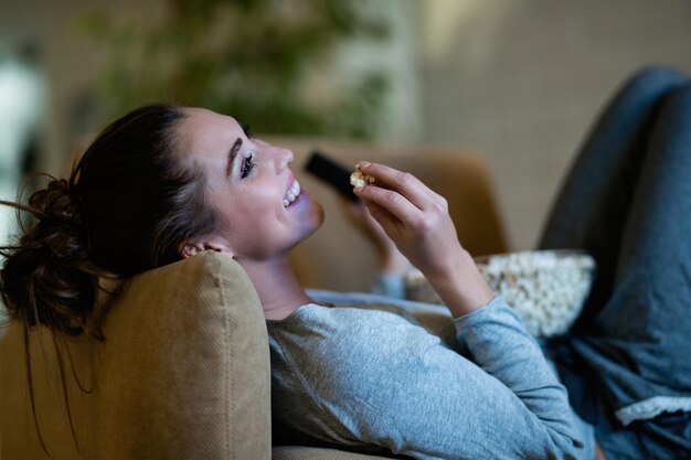 Jeune femme heureuse se détendre dans un fauteuil la nuit et regarder un film en mangeant du pop-corn