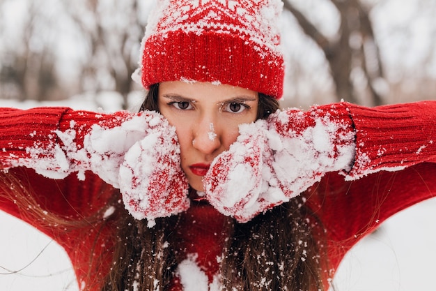 Photo gratuite jeune femme heureuse souriante assez candide dans des mitaines rouges et un chapeau portant un chandail tricoté marchant jouant dans le parc dans la neige, des vêtements chauds, s'amuser, une grimace sérieuse