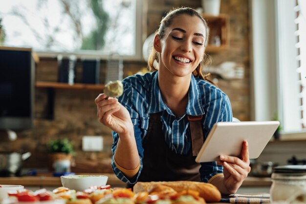 Jeune femme heureuse utilisant une tablette numérique tout en faisant de la bruschetta dans la cuisine.