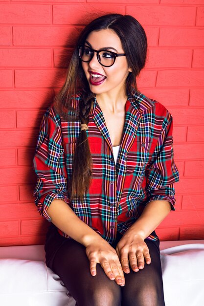 Jeune femme hipster assez séduisante avec de longues jambes s'amusant et assise sur un canapé blanc, portant une chemise à carreaux et des lunettes claires, des cheveux longs et un maquillage lumineux.