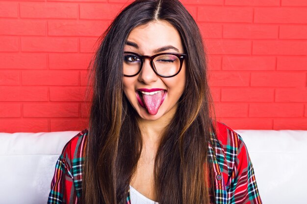 Jeune femme hipster assez séduisante s'amusant et montrant une longue langue et un clin d'œil, portant une chemise à carreaux et des lunettes claires, des cheveux longs et un maquillage lumineux. Mur urbain rouge.