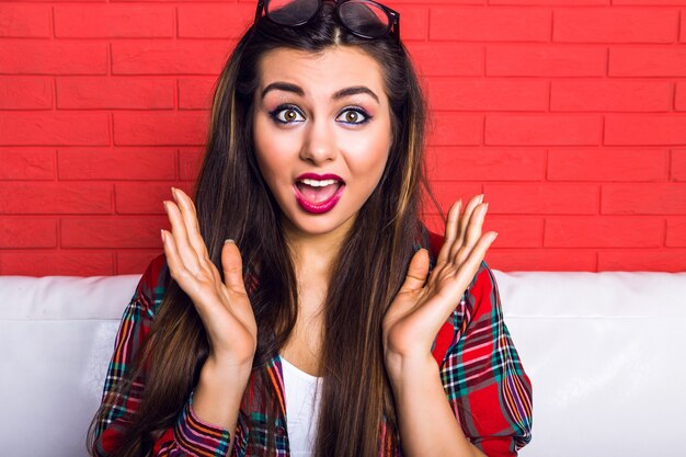 Jeune femme hipster assez séduisante s'amusant et montrant une longue langue et un clin d'œil, portant une chemise à carreaux et des lunettes claires, des cheveux longs et un maquillage lumineux.