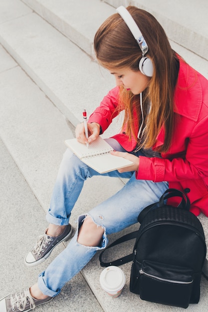 Jeune femme hipster en manteau rose, jeans assis dans la rue avec sac à dos et café en écoutant de la musique au casque, étudiant prenant des notes