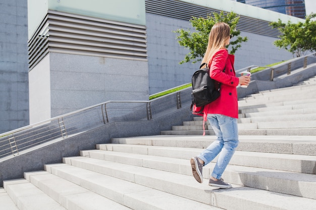 Jeune femme hipster en manteau rose, jeans marchant dans les escaliers dans la rue avec sac à dos et café en écoutant de la musique sur des écouteurs, portant des lunettes de soleil