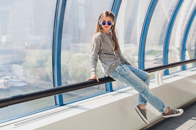 Jeune femme hipster en tenue décontractée s'amusant à écouter de la musique dans les écouteurs, portant des jeans, pull et lunettes de soleil, style urbain