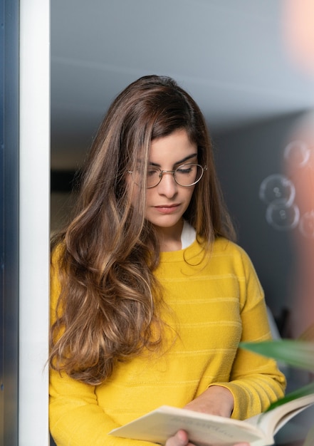 Jeune femme hispanique dans une chemise jaune debout près de la fenêtre et lisant un livre