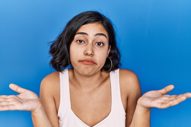 Jeune femme hispanique debout sur fond bleu expression désemparée et confuse avec les bras et les mains levés. notion de doute.