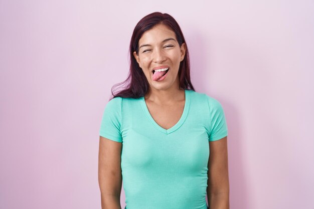 Jeune femme hispanique debout sur fond rose tirant la langue heureuse avec une drôle d'expression. notion d'émotion.