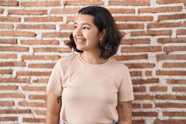 Jeune femme hispanique debout sur un mur de briques regardant de côté avec un sourire sur le visage expression naturelle rire confiant