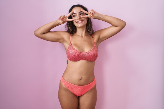 Jeune femme hispanique portant de la lingerie sur fond rose faisant le symbole de la paix avec les doigts sur le visage, souriant joyeusement montrant la victoire