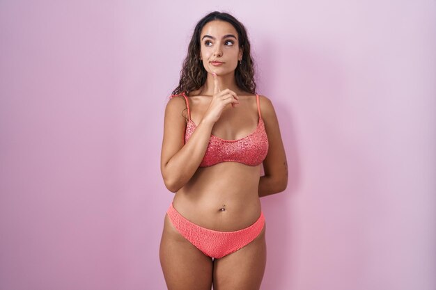 Jeune femme hispanique portant de la lingerie sur fond rose pensant concentrée sur le doute avec le doigt sur le menton et levant les yeux se demandant