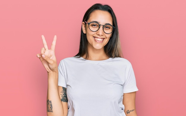 Jeune femme hispanique portant un t-shirt blanc décontracté souriant regardant la caméra montrant les doigts faisant le signe de la victoire. numéro deux.