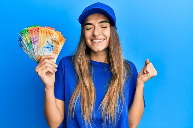 Jeune femme hispanique portant l'uniforme de livraison et la casquette tenant le franc suisse criant fier de célébrer la victoire et le succès très excité avec le bras levé