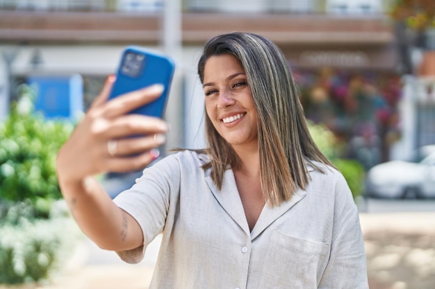 Une jeune femme hispanique souriante et confiante se fait un selfie avec son smartphone dans la rue.