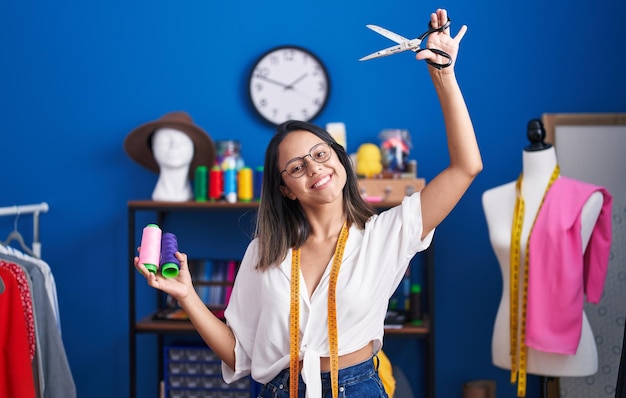 Jeune femme hispanique tailleur souriant confiant tenant des ciseaux et du fil au studio de couture